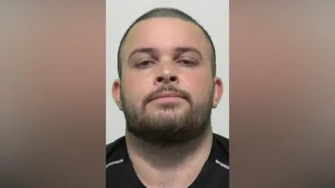 Northumbria Police Mugshot of Roberts. He has a large round head with shaved black hair and a black beard. 