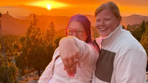 Hayley Cook A picture of Hayley on the left and Lizzie on the right, Hayley is holding out her left hand to show off her engagement ring, there are trees and mountains and the sun setting behind them.
