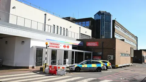 The front of a hospital building on a sunny day. A couple of police cars are parked outside. 