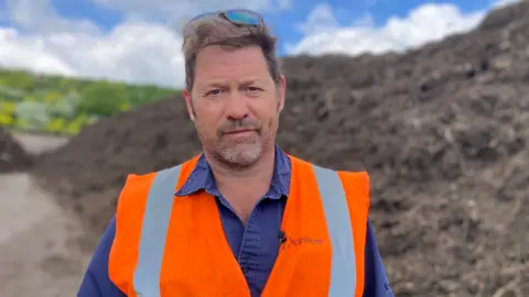 Fiona Irving / BBC A white man with brown hair and a short beard and moustache looks at the camera. He is wearing a blue collared shirt and an orange high-vis vest. He has sunglasses perched on his head. In the background is a large bank of brown compost.