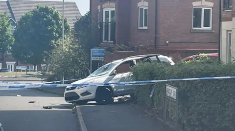 BBC Blue and white police tape is in the foreground, near to an Arbour Drive road sign by a hedge on the right on the photo. A white car is in the centre of the image, with buildings in the background.