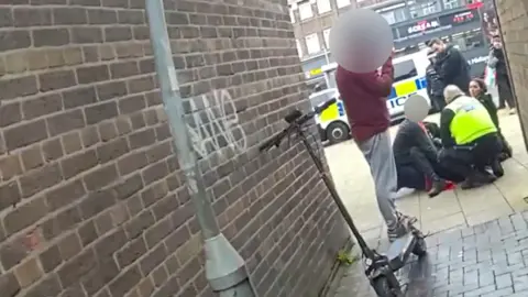 An image at an angle - presumably because it is from a police body worn camera. It shows a brick alleyway with a black e-scooter leaning up against the wall next to a grey lamppost. A young person is standing next to it with their face blurred entirely. They are wearing a red top and grey tracksuit bottoms. Beyond him is a group of police officers on the floor around another officer, who only has one leg visible.