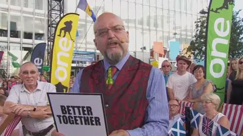 A middle aged bald man in glasses, with a grey goatee addresses the camera, while a crowd of people watch behind him. He is wearing a red floral patterned waistcoat, a red- and yellow- tartan tie. He is holding a card with the words 'Better Together' printed on the back.