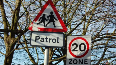 BBC A red triangle "Patrol" sign with schoolchildren pictured and a round 20mph road sign denoting a 20mph zone