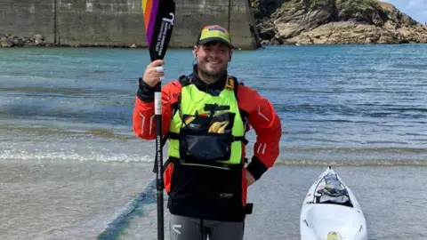 A man wearing a yellow life jacket, wearing a red hat and holding an oar on a beach