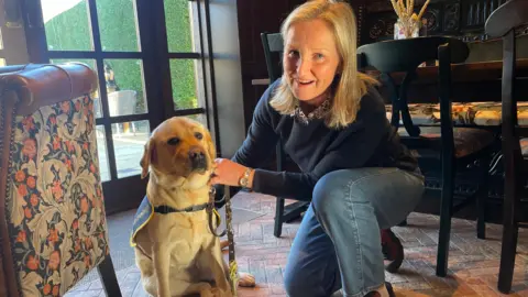 BBC/Jack Haddaway-Weller A woman kneels on one knee next to her Labrador puppy. She is wearing a navy blue jumper and blue jeans and is smiling.