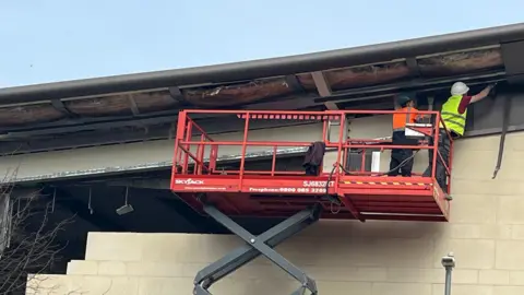 Two men in high-vis tops and wearing safety helmets stand on a cherry-picker by a wall which is being knocked down