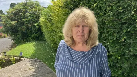 Ms Bannister pictured in her garden wearing a blue top with white vertical stripes. She has blonde shoulder-length wavy hair with a fringe. 
