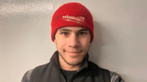 Kelechi Agoha wears a red woollen had which reads 'Milwaukee' in white wont on the front of it. He has short brown stubble along his chin and cheeks. He wears a black gillet and black and grey camo jumper beneath it. He is looking directly into the camera.