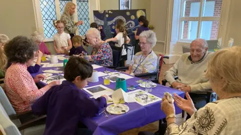 Helen Mulroy/BBC Residents and pupils can be seen at Margaret House care home sat at a table taking part in a Vincent Van Gough art project. 