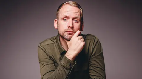 Matthew Gilmore Andi looks at the camera with an inquisitive expression, resting his chin on one hand. He has short blond hair, a stubbly beard and wears a long sleeve dark green top. 