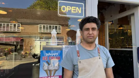 Jo Burn/BBC A man in a blue t-shirt and a beige apron standing outside a shop window. There is a neon open sign and an advert for ice cream in the window.