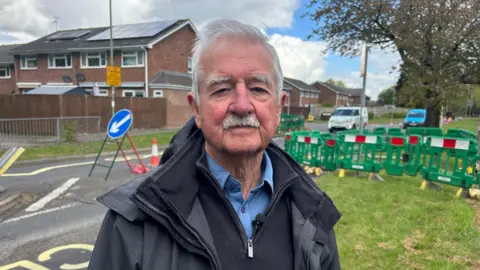 Bob Lloyd stands near the roadworks. He has white hair and a moustache and wears a dark anorak.