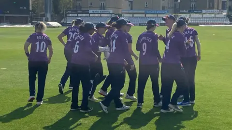 James Patient/BBC The Essex Women's team wearing purple tops and black trousers. They are standing on the pitch ahead of a game.