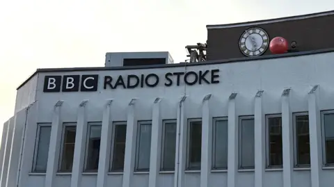 BBC Radio Stoke's building, a white building with a series of windows, with a clock on top. Presenter Lee Blakeman is stood by the clock, holding a large red medicine ball.