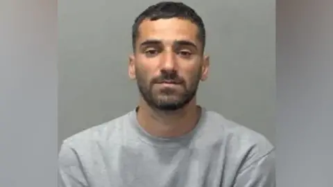 Barzel Faesal in a grey sweatshirt looks directly at the camera as he is photographed for a police mugshot. 