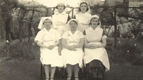 Family photo Five nurses seen in black and white, with three of them seated in front and two stood behind. They are wearing an old fashioned nurses uniform from the 1940s with white hats.
