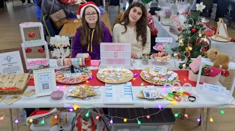 Family A girl with long brown hair, glasses, a santa hat and a purple hoodie and a girl with long brown hair and a cream jumper - sit behind a table with a white table cloth and plates with bracelets. A light box says Beaded Beauty.