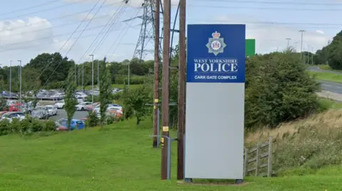 A large free-standing sign placed on a grassy area of land. The sign is grey with the upper section in blue with white with the words West Yorkshire Police Carr Gate Complex written on it. In the background are parked cars in a car park.