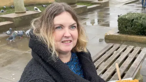 Robby West/BBC Sharon Williams is sitting on a bench on the left. She has shoulder length blonde hair and is wearing a black coat and blue and black top. Behind her are pigeons eating off a walkway. 