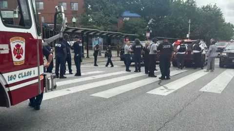 A landscape image of a road crossing with multiple emergency service workers standing around. The front of a red fire truck can be seen on the left hand side.