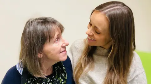 Patient Samantha Dennison is sitting on a hospital bed and looking at her daughter Steph. Samantha has shoulder length brown hair and wears a dark blue cardigan over a floral top. Steph has long brown hair and wears a light grey,  high-neck jumper.