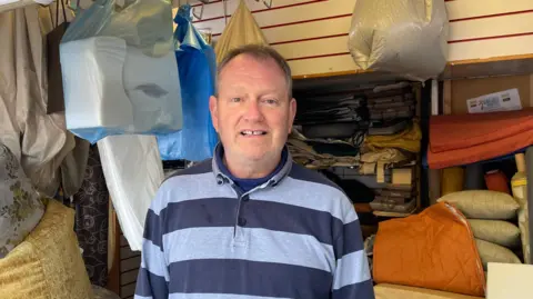 LDRS Steve Taylor wearing a striped blue and grey polo shirt stood in a room in his shop surrounded by cushions and bags of foam material.