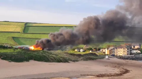 A beachside building is on fire with large plumes of black smoke coming away from it and over the beach. Fire crews are seen spraying the property with water. 