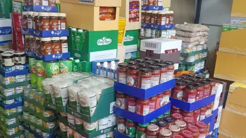 Luton Foodbank Large amounts of tinned and jarred food, all in boxes, piled in a room. 