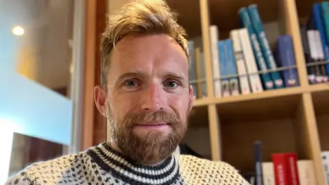 A writer with dark blond hair and beard, wearing a grey and white jumper.