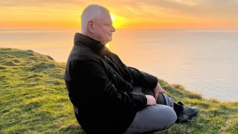 Diocese of Sodor and Man Andrew Hillas sitting on Peel Hill. with the sun setting behind. He has short grey hair and is wearing a black jacket and grey trousers. The sea stretches off the horizon behind him and the setting sun illuminated the grass around him.