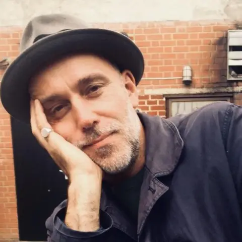 Jamie Thraves A man smiles at the camera, with his head to one side, learning on his hand, he has a hat on and is standing in front of a brick wall.