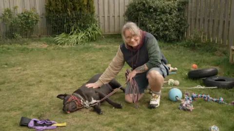 Nina Morgan kneels on the grass in a garden, gently resting a hand on a large black dog named Frankie lying on its side. Frankie is wearing a red head collar and leash. Various dog toys, including ropes and balls, are scattered around the garden, which is enclosed by wooden fencing and greenery.