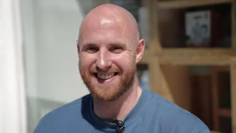 Euan Spark, a bald man with a ginger beard, smiles at the camera. He is standing outside a cafe.