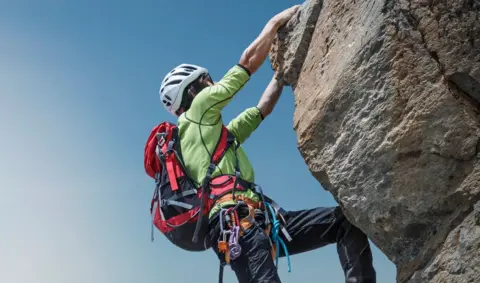 Stock image of a female climber wearing a backpack hanging onto a rock