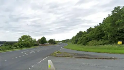 A dual carriageway at a junction on a cloudy day. Trees lines the road on either side. A red brick building can be seen in the background on the left.