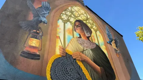 A colourful, large-scale wall painting showing a woman dressed in modest 19th Century clothing while knitting a woollen jumper. She is depicted standing in front of an arched church window through which light streams. To the left, a black bird carries a lamp. Another bird flies to the right.