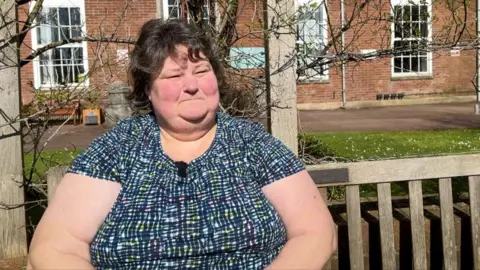 A woman sits on a bench in the sunshine outside a community hospital at Torbay. She has short brown hair, and is wearing a short sleeved blue checked top. 