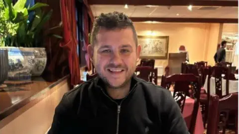 A man sits at a restaurant table facing the camera, wearing a dark zip-neck top. The restaurant has wooden furniture, red curtains, warm lighting and a large plant beside the seating area.
