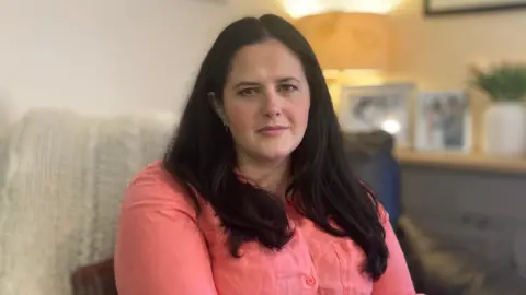 BBC Claire Sugden, former justice minister, sitting on a couch and looking at the camera. She is wearing a salmon colour blouse. 