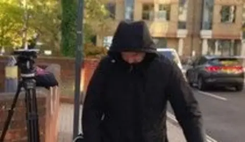 Gregory Assemakis: a man looking down and wearing a black anorak with the hood up. To his right is a wall and a camera tripod. In the background is a building and black car.