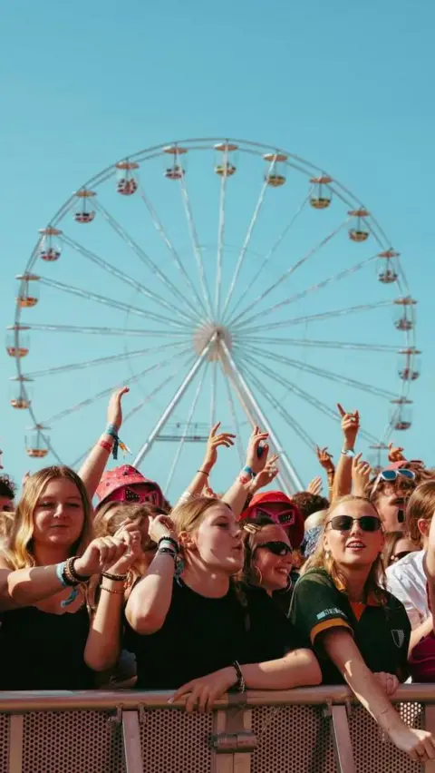A crowd of festivalgoers stands at the front barrier with arms raised while a large Ferris wheel rises in the background against a clear blue sky.