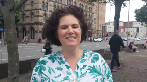 Claire Hanna. She has short brown curly hair, wearing a green floral blouse. She has a rainbow on her cheek.