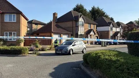 A street in a housing estate with detached houses. There is a line of blue and white police tape stretched across the road and a car immediately behind it.