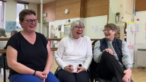 Three women, all wearing glasses sitting and laughing in a dance studio. The woman in the middle is sat with a cup of tea and wearing a long sleeve white top. The woman to the right of her, is wearing a grey jumper and a dark blue gilet. And the woman on the left is wearing a black t-shirt and has her hands crossed on her lap.