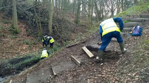 Friends of Holywell Dene Four volunteers are working to install new steps up a hill in woodland. One person in a hi-vis jacket is working on the steps while three others are in the background working near a stream.