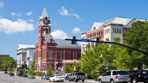 Getty Images A street view of eleven cars travelling through traffic lights on the right is three buildings. The building furthest away is white, the second is a red brick town hall and the closest is a building made of white and red brick.