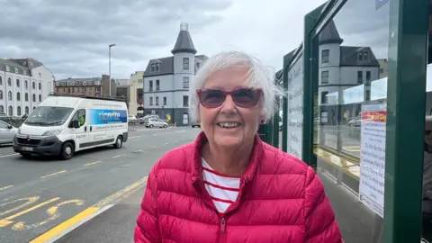 By the side of a road, and in front of a bus stop, Beryl Payton stands in a red jacket, with a white and red striped shirt. She is smiling whilst wearing red rimmed sunglasses. 