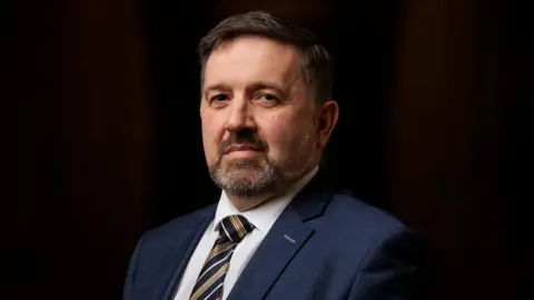 PA Media Robin Swann looking straight to camera, wearing a blue suit white shirt and diagonally striped gold and blue tie. He has black hair and a beard.