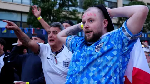 Owen Humphreys/PA A man with the word "England" painted across his forehead stares intensely at the game with his hands behind his head. Behind him, a man who appears somewhat agitated points at the screen.